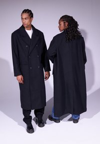 Manteau noir croisé en tissu épais, avec des poches latérales et une silhouette droite, porté par deux personnes de dos.