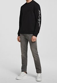 Hombre vestido con una camiseta de manga larga negra con un texto blanco en la manga, jeans grises y zapatillas blancas, de pie frente a un fondo liso.