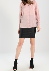 Helles rosa Langarm-Bluse mit V-Ausschnitt und Knopfverschluss, kombiniert mit einem taillierten schwarzen Kunstlederrock und offenen schwarzen Stiefeln.