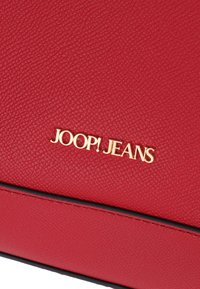 Rdeča teksturirana usnjena torba z zlatim logotipom "JOOP! JEANS" in kontrastnim črnim šivom ob spodnjem šivu.