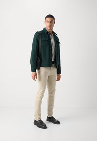 Veste vert foncé en tissu texturé, deux poches poitrine, boutons marron, chemise crème, pantalon beige et chaussures noires avec des accessoires en or.