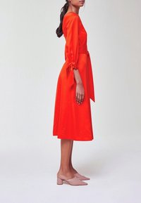 Femme portant une robe midi rouge vif à manches trois-quarts nouées aux poignets, debout de profil sur un fond clair uni.