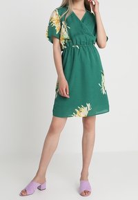 Robe verte à manches courtes et enroulée avec un motif floral, taille cintrée et forme évasée. Associée à des mules à talons block lilas.