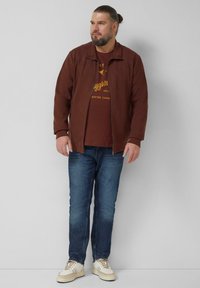 Brązowa kurtka ze sztruksu z zapięciem na zamek, noszona na bordowej koszulce z nadrukiem, zestawiona z niebieskimi dżinsami i kremowymi sneakersami. Gładkie tło.