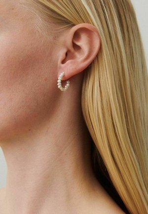 Približni prikaz ženinog uha s ravnom plavom kosom koja nosi malu obručastu naušnicu od bijelih bisera.