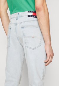 Jeans de mezclilla de color azul claro con parche de logo de Tommy Jeans en la cintura y pequeño emblema de bandera en el bolsillo trasero, llevados con una blusa verde.