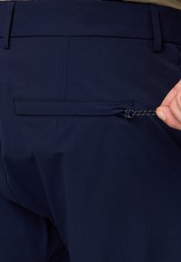 Tamno plave hlače s stražnjim džepom sa zatvaračem. Tkanina je glatka s krojem koji ističe minimalne detalje šivanja.