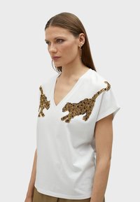 T-shirt bianco a V con due design di leopardo ricamati in oro e nero sul davanti, maniche corte e una vestibilità morbida.