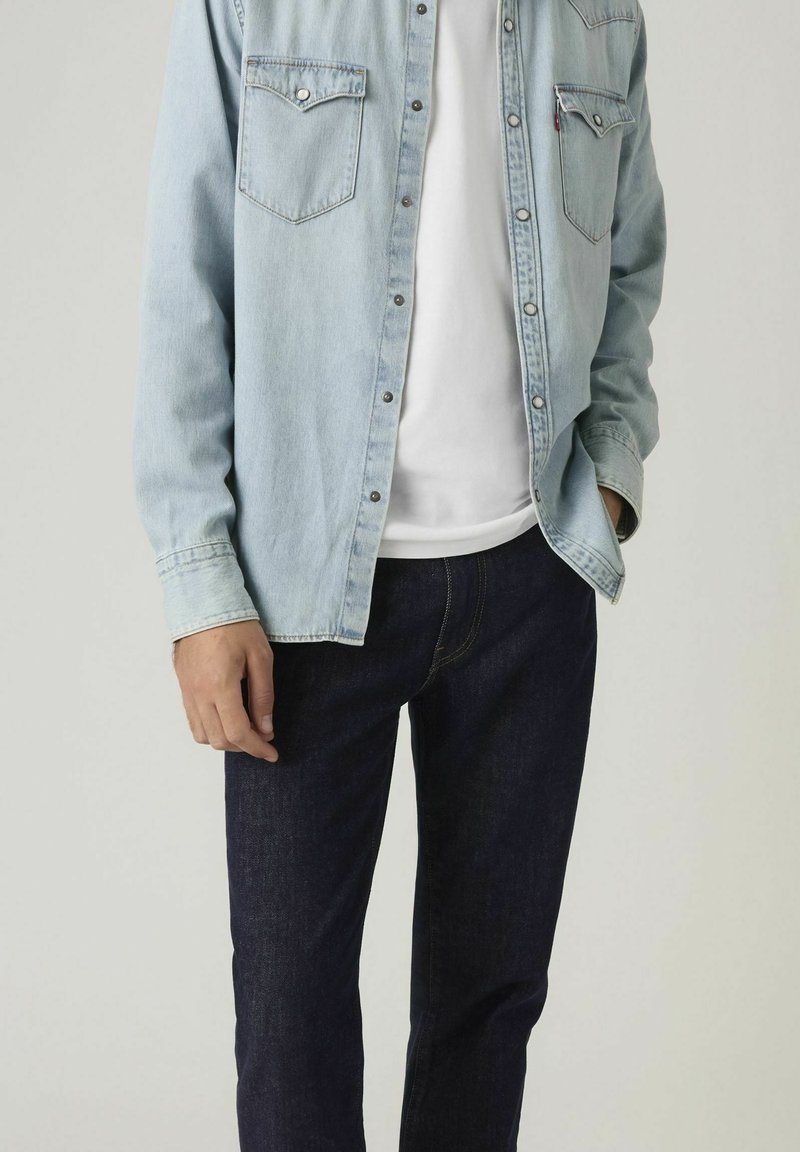 Uomo con camicia di jeans azzurro chiaro sopra una t-shirt bianca e jeans blu scuro, mano in tasca, su sfondo neutro.