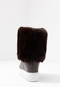 Bota de tacón bajo de piel sintética marrón con un puño alto, cuerpo similar al cuero y suela de goma blanca texturizada. Presenta un logo en la suela.