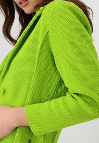 Cárdigan de punto verde neón con tejido texturizado, que cuenta con un cierre de botones y una silueta ajustada. Mangas largas y costuras detalladas visibles.