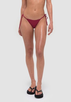 Kobieta w bordowych bikini z wiązaniami po bokach i czarnych klapkach na platformie, stojąca na jasnym, jednolitym tle.