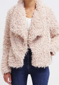 Fuzzy, ljusrosa jacka med sjalkrag, öppen front och mjuk textur. Har långa ärmar och en lös passform, kombinerad med mörka jeans.