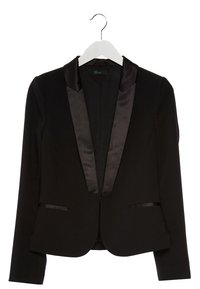 Svart kavaj med satinnotchkragar, två framfickor och strukturerade axlar. Texturerad tyg med en slät yta. Hänger från en vit krok.