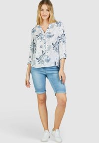 Leichtes, blumenmusterndes Blusenshirt in Weiß und Blau, mit dreiviertel Ärmeln und V-Ausschnitt; kombiniert mit hellblauen, gekrempelten Shorts und weißen Sneakern.