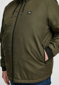 Jachetă padding verde măslin cu fermoar negru, manșete elastice și buzunare laterale. Prezintă o aplicație cu logo pe zona stângă a pieptului.