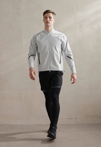 Lys grå treningsjakke med svarte detaljer, glidelås foran, kombinert med svarte shorts og tettsittende leggings, båret av en mann som går.
