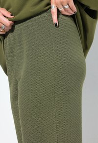 Close-up van een persoon in gestructureerde olijfgroene broek, met een hand die een shirt omhoog trekt en naar beneden wijst, terwijl zilveren ringen en zwarte nagellak worden getoond.