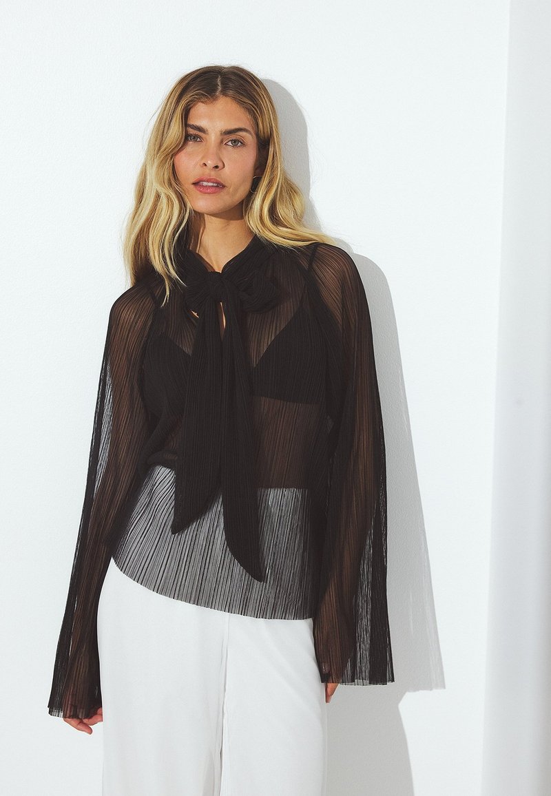 Blusa nera a pieghe con scollo a cravatta, maniche lunghe trasparenti e design a strati su una base grigia a contrasto. Texture liscia, tessuto leggero.