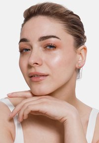 Goldene Ohrstecker mit kreisförmigem Design und glatter Oberfläche. Das Modell hat helle Haut, einen natürlichen Makeup-Look und hält die Hand unter das Kinn.