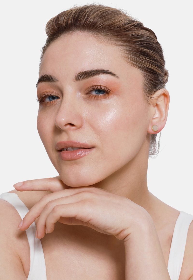 Goldene Ohrstecker mit kreisförmigem Design und glatter Oberfläche. Das Modell hat helle Haut, einen natürlichen Makeup-Look und hält die Hand unter das Kinn.