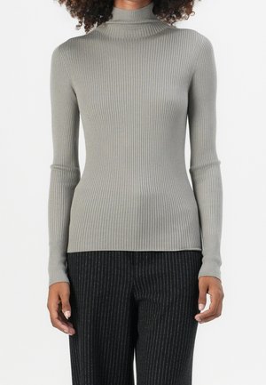 Personne portant un pull gris côtelé ajusté à col roulé et un pantalon noir à fines rayures sur un fond uni.