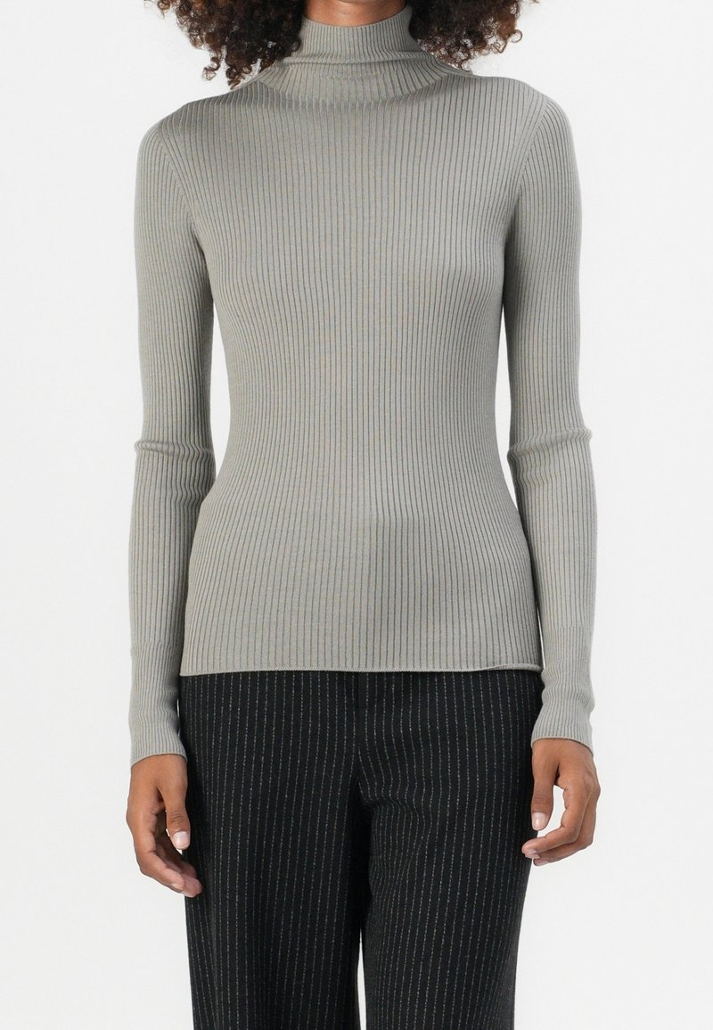 Personne portant un pull gris côtelé ajusté à col roulé et un pantalon noir à fines rayures sur un fond uni.