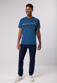 Tyrkysové bavlněné tričko s bílými horizontálními pruhy přes hruď a malým textovým detailem, kombinované s tmavými denimovými džínami a bílými teniskami.