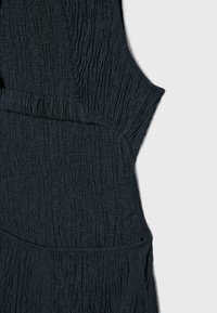 Gros plan sur un tissu texturé foncé avec un détail de couture froncée, montrant une section de vêtement sans manches sur un fond clair uni.