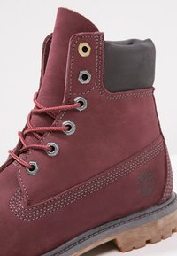 Burgundy leather boot featuring a black padded collar, metal eyelets, red laces, and a textured brown rubber sole with a logo detail.