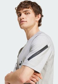 Šedé tričko s krátkým rukávem z měkké látky, s černými akcenty na rukávech a jemným vzorem na přední straně.