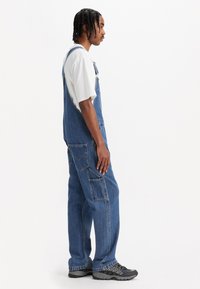 Salopette en denim bleu moyen avec deux poches latérales et des bretelles réglables, associée à un t-shirt blanc et des chaussures de randonnée noires.