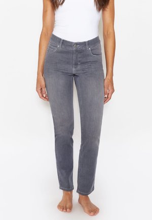 Frau trägt eng anliegende graue Jeans und ein weißes ärmelloses Oberteil, steht barfuß vor einem schlichten weißen Hintergrund.