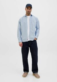 Camicia azzurra chiara con bottoni, maglietta bianca, pantaloni di denim scuri, scarpe beige senza lacci. La camicia presenta una tasca e un dettaglio rosso.