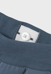 Grijze geribbelde tailleband met een witte stoffen patch en een transparante knop met een viergaatjesontwerp. Gladde textuur en stevig materiaal.