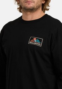 Uomo che indossa una maglietta nera a maniche lunghe con una piccola patch del logo Billabong che presenta montagne, un'onda e il sole sul lato sinistro del petto.