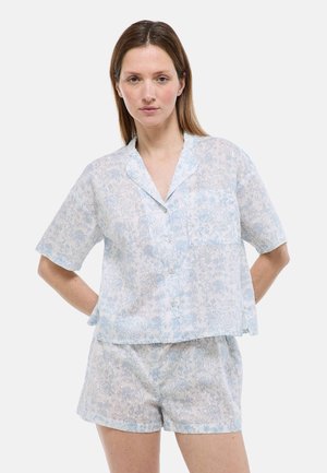 Femme portant une chemise à manches courtes boutonnée à fleurs bleu clair et blanc avec un short ample assorti, debout avec les mains derrière le dos.