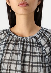 Blusa a quadri grigia con scollo arricciato e dettagli con bottoni, realizzata in tessuto leggero e textured. Presenta orecchini a goccia delicati.