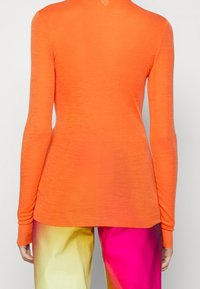 Langärmliges Oberteil aus leuchtend orangefarbenem Stoff, mit weicher Textur und tailliertem Schnitt, kombiniert mit mehrfarbigen, glänzenden Hosen mit weitem Bein.