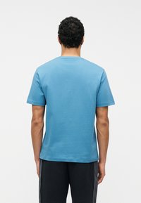 Άνδρας με σγουρά μαλλιά που φοράει απλό γαλάζιο κοντομάνικο πουκάμισο και μαύρη βερμούδα στέκεται γυρισμένος με την πλάτη προς έναν απλό λευκό φόντο.