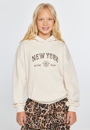 Blondynka ubrana w kremową bluzę z kapturem New York i brązową plisowaną spódnicę w cętki lamparta, stojąca na jednolitym tle, uśmiechająca się.