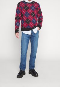 Pull à motif argyle rouge, bleu et noir avec un col rond et des poignets côtelés, porté sur une chemise blanche à col, associé à un jean bleu et des chaussures noires.