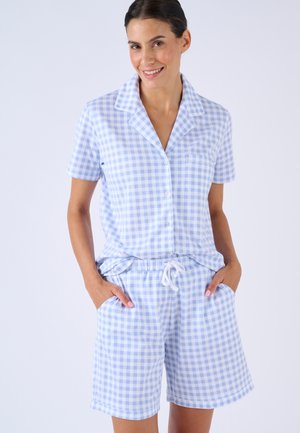 Vrouw lacht, draagt een lichtblauw-wit geruit overhemd met korte mouwen en bijpassende short met trekkoord, handen in de zakken.