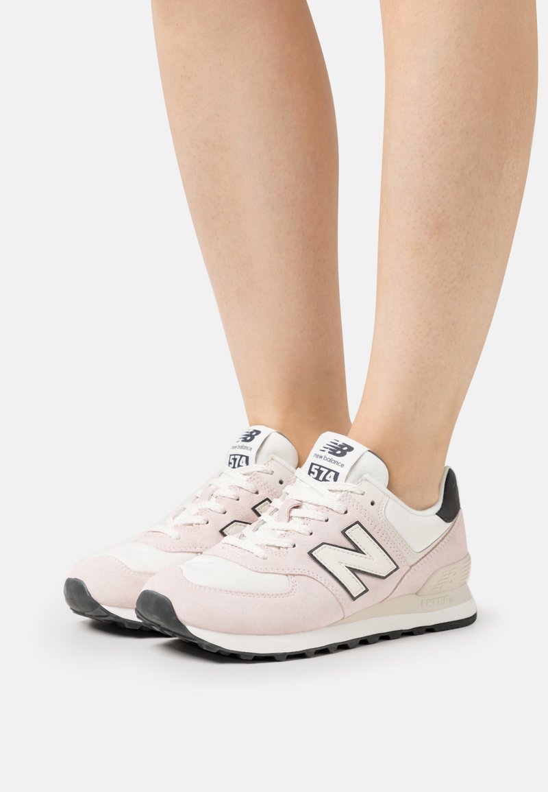 Paire de baskets New Balance 574 rose clair et blanc portées sur des jambes nues avec une semelle noire et des détails du logo.