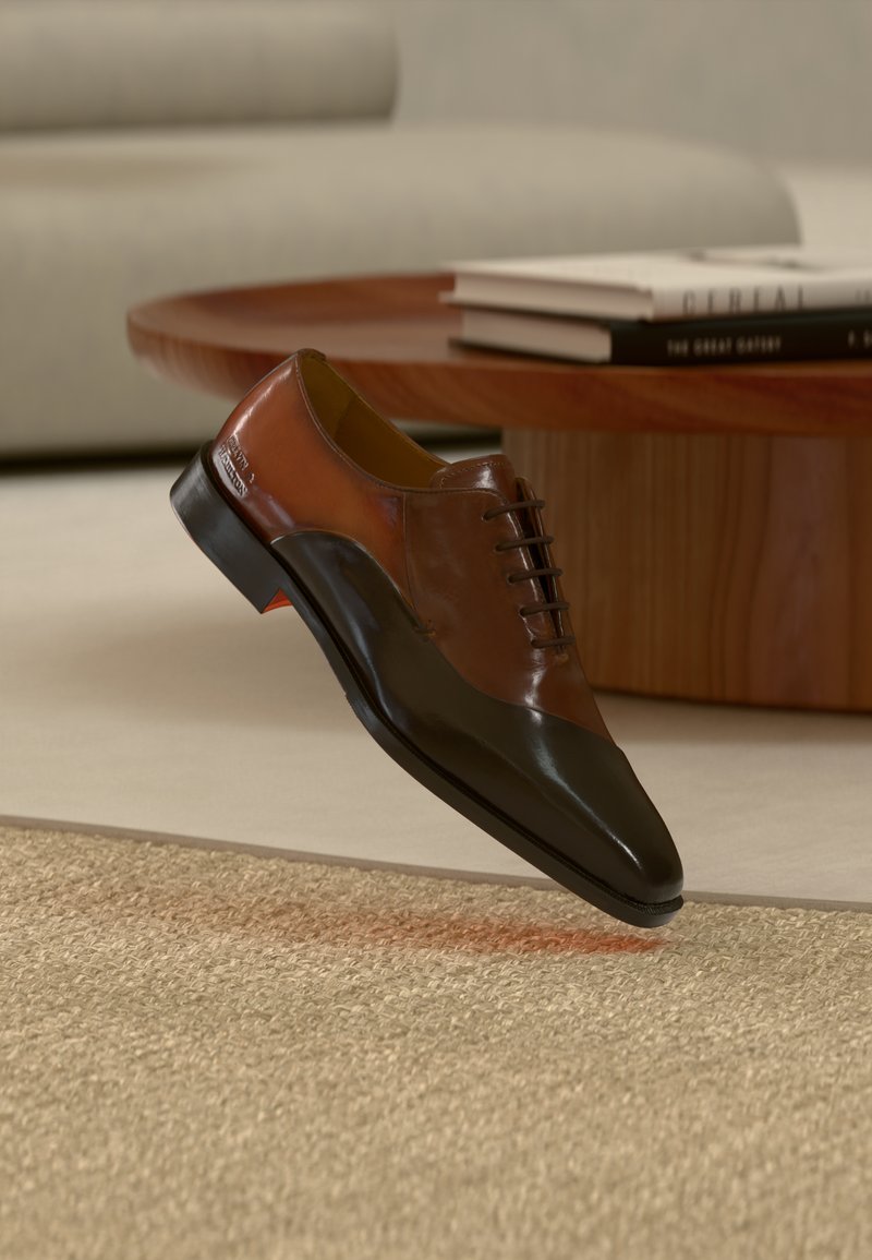 Chaussure habillée en cuir marron et noir flottant au-dessus d'un tapis beige texturé, avec une table basse en bois et des livres empilés en arrière-plan.