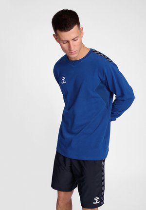 Maglia sportiva a maniche lunghe blu con scollatura a V, dotata di pannelli laterali testurizzati a contrasto e accenti a chevron neri sulle spalle. Pantaloni corti neri.