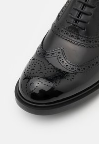 Chaussure brogue en cuir noir brillant avec perforations décoratives et laçage à l'avant, vue en gros plan sous un angle plongeant.