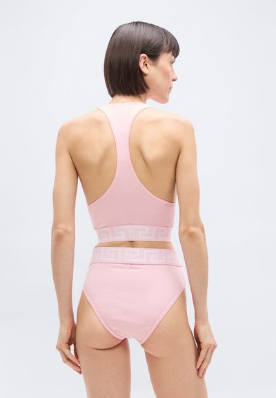 Γυναίκα με κοντά καστανά μαλλιά που φοράει ένα ανοιχτό ροζ crop top με racerback και αντιστοιχούσα ψηλόμεση εσώρουχο με λευκό γεωμετρικό σχέδιο στη μέση.