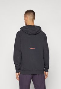 Mørkegrå hoodie med kengurulomme og snøre på hætten. Har Reebok-logo i rød på ryggen, lavet af blødt, tekstureret stof.