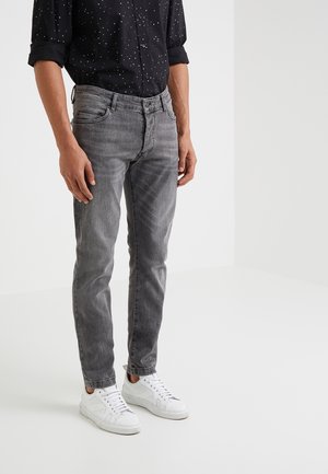 Man wearing rolled-up sleeves black shirt with white dots, gray slim-fit jeans, and white sneakers standing against a plain background.